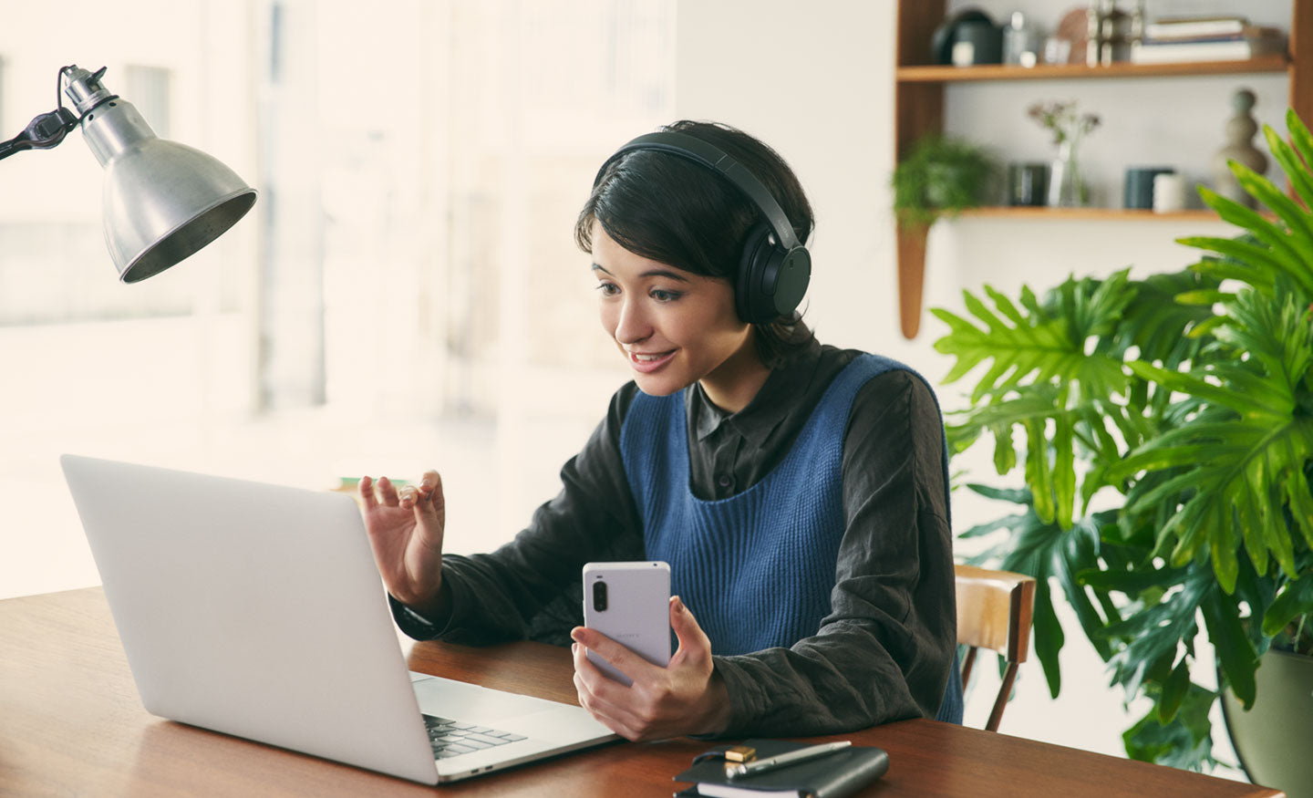 Image of a woman at a desk wearing a black pair of Sony WH-CH720 headphones and using a laptop and mobile phone